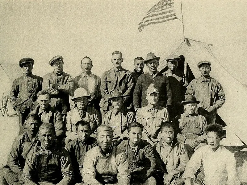 An old black and white photo of a large group of men in hats and field clothes posed outside a tent
