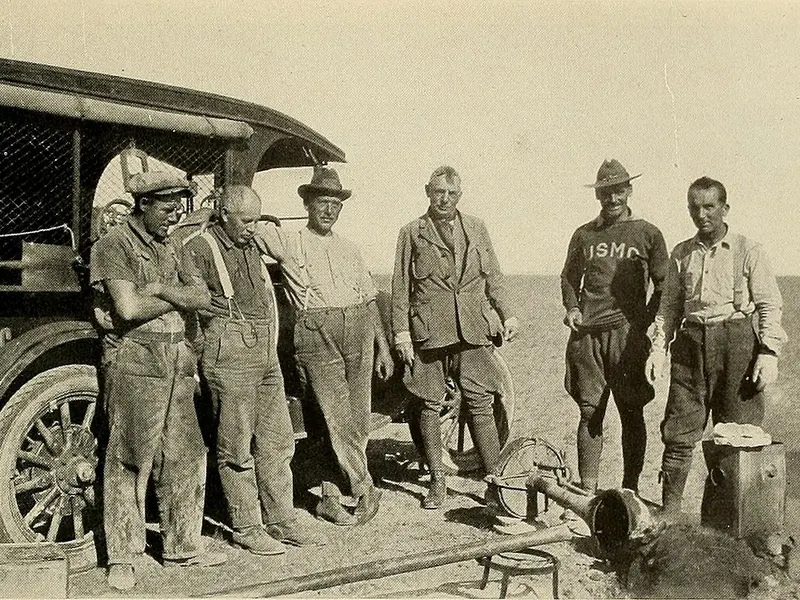 An old black and white photo of men repairing an early motor car on open grassland